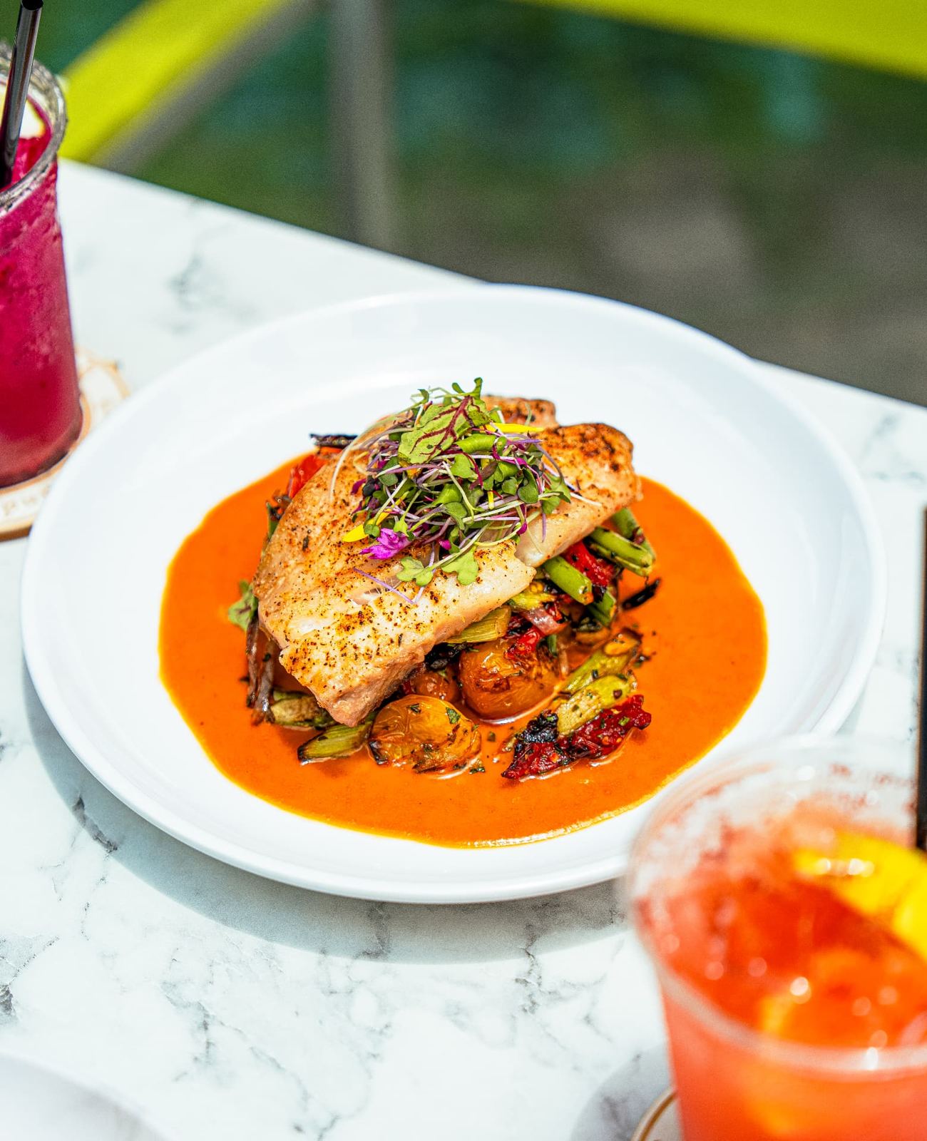 Mahi-mahi plated with roasted vegetables beside a red cocktail